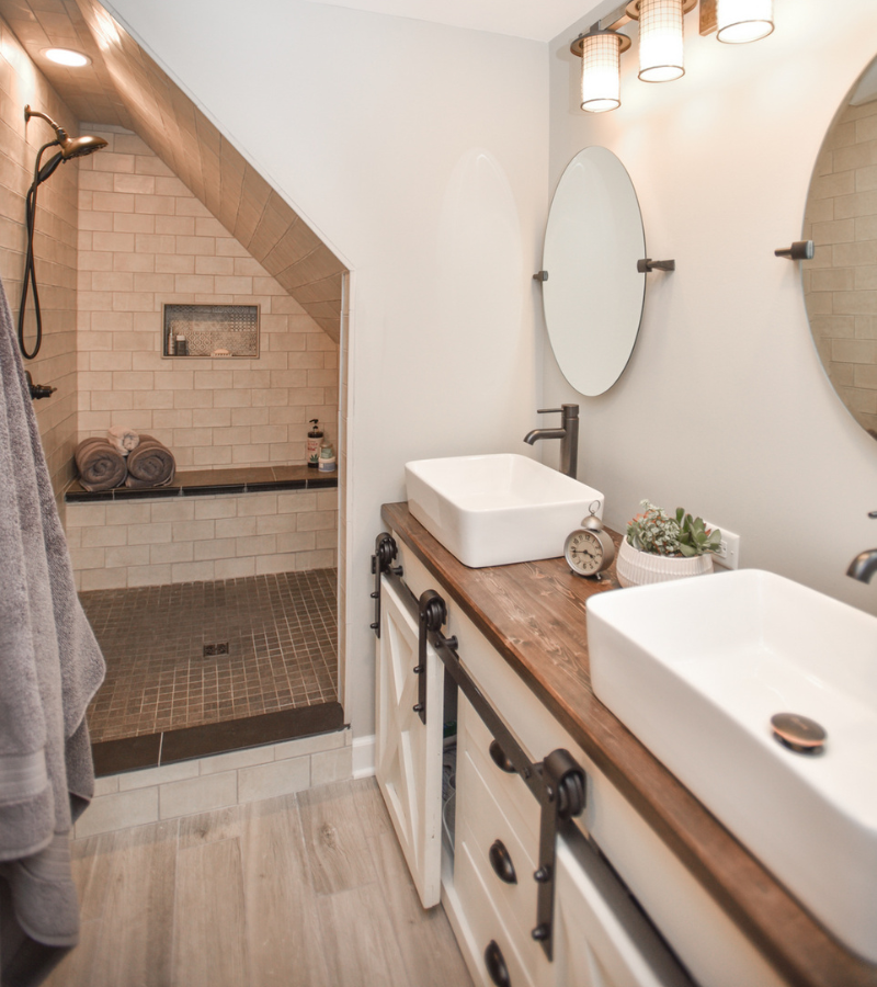 Rustic bathroom with double vanity, vessel sinks, sliding barn door cabinetry, and walk-in shower under angled ceiling.